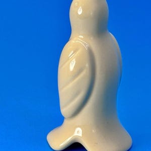 May include: A ceramic bird figurine in a light beige color. The bird has a rounded body, a small head with an open beak, and detailed wing features. The figurine is set against a solid blue background.