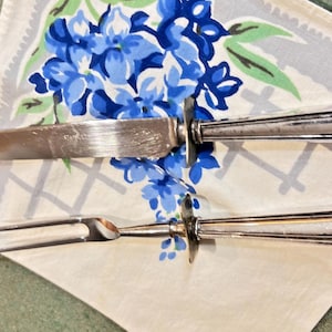 May include: A silver-toned carving knife and a two-pronged carving fork with rectangular handles, resting on a white fabric napkin featuring a blue floral pattern with green leaves and a grey grid design printed on the textile surface.