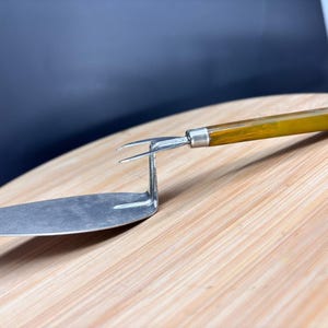 May include: A vintage silver-toned pie server with a forked end and a long, flat blade. The handle is a warm amber color. The server is resting on a light-colored wooden surface.