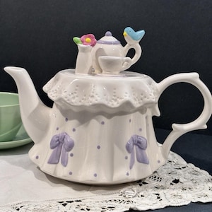 May include: A white ceramic teapot featuring a ruffled lace-like rim, purple polka dots, and raised purple bows. The lid is adorned with a miniature teapot, a small teacup, a vase with a pink flower, and a small blue bird perched on the handle.