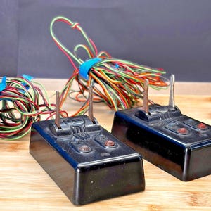 May include: Two black vintage electrical components with red and green wires. Each component has two metal prongs and two red buttons. Bundles of colorful wires are secured with blue tape.
