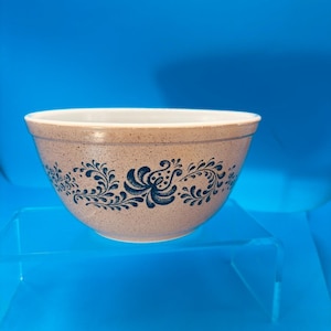 May include: A beige stoneware mixing bowl with a speckled finish. The bowl features a blue floral design around the exterior. The bowl is sitting on a clear, angled surface against a blue background. The bowl is a kitchenware item.