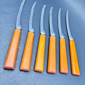 May include: Set of six vintage fruit knives. Each knife features a curved stainless steel blade and a bright orange handle. The handles have a rectangular shape with a slightly tapered design. The knives are arranged in a row.