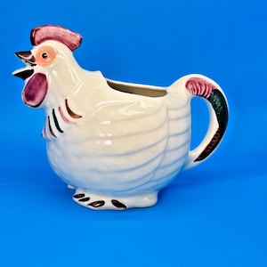 May include: A white ceramic rooster creamer with a red comb and wattle, black and brown accents, and a handle. The creamer is designed to resemble a rooster and is set against a blue background.