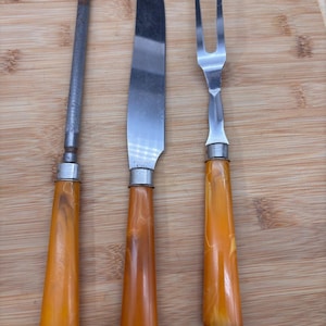May include: A knife sharpening steel, a carving knife, and a carving fork set on a wooden cutting board. The handles are a marbled orange color. The knife blade is stainless steel.
