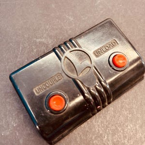 May include: Black vintage Lionel train accessory with two red buttons and the text "UNCOUPLE" and "UNLOAD". The accessory has the Lionel logo in the center.