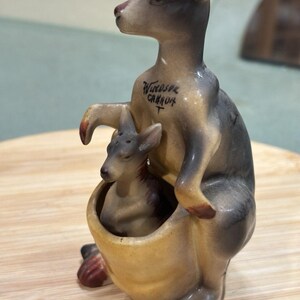 May include: A ceramic kangaroo figurine with a smaller joey inside its pouch. The kangaroo is painted in shades of tan, brown, and grey. The text Windsor Canada is printed in black on the chest of the larger kangaroo. The figurine has small holes on the top of both heads.