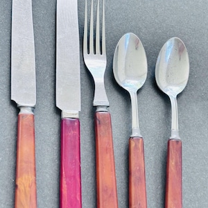 May include: A set of five pieces of flatware including two knives, one fork, and two spoons. Each utensil features a metallic silver head and a rectangular handle with a marbled, reddish-brown and orange resin finish.