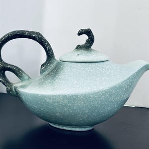 May include: A ceramic teapot with a light blue and white speckled glaze. The teapot features a dark, textured, organic-shaped handle and a matching lid with a small, curved finial. The body has a wide, rounded base and a tapered spout.