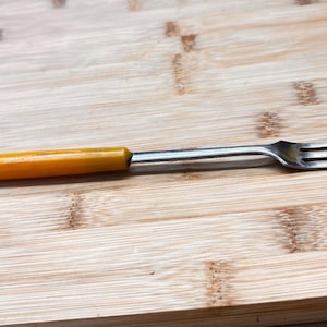 May include: A vintage fork with a stainless steel head and a yellow handle. The fork is lying on a wooden cutting board. The handle is a solid, warm yellow color. The tines are thin and sharp.