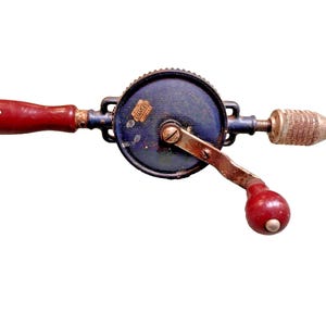 May include: A vintage hand drill with a blue metal body, a red wooden handle, and a brass chuck. The drill has a crank handle and a metal gear.