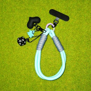 May include: A light blue phone lanyard with gray accents and a black phone grip. The lanyard features a black heart charm, a blue ghost charm, and a black dice charm. The background is a solid green.