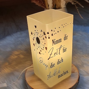 Könnte beinhalten: Eine weiße, gefrostete Glaslampe mit einem Löwenzahnmuster und dem Text "Nimm dir Zeit für Dinge, die dich glücklich machen." Die Lampe steht auf einem Holzsockel und ist beleuchtet.