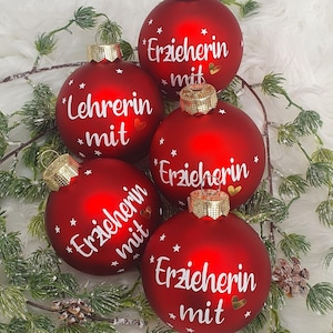 Könnte beinhalten: Fünf rote Weihnachtskugeln mit goldenen Kappen und weißem Text. Die Kugeln tragen die Wörter "Erzieherin mit" und "Lehrerin mit", sowie kleine weiße Sterne und goldene Herzen. Die Kugeln sind von grünen Zweigen umgeben.