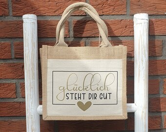 May include: A small, brown burlap tote bag with a white canvas panel. The panel has a gold heart and the text "glücklich steht dir gut" in gold lettering.