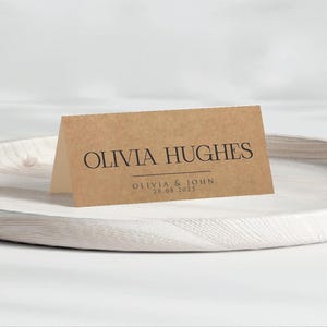 Può includere: Un segnaposto piegato, color beige, con il nome "OLIVIA HUGHES" stampato in nero, centrato. Sotto, un testo più piccolo recita "OLIVIA & JOHN 28.08.2025". Il biglietto è su un vassoio rotondo di legno bianco.