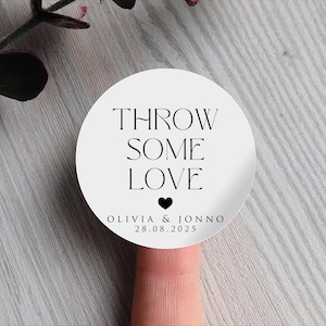 Könnte beinhalten: Ein runder, weißer Aufkleber mit dem schwarzen Text "THROW SOME LOVE", einem schwarzen Herz und den Namen Olivia & Jonno mit dem Datum 28.08.2025. Der Aufkleber befindet sich auf einer hellen Holzoberfläche.