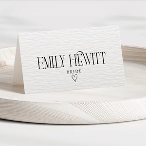 Pu&ograve; includere: Un biglietto bianco strutturato con il nome "EMILY HEWITT" in corsivo nero, con "BRIDE" sotto e un simbolo a forma di cuore. Il biglietto &egrave; esposto su un vassoio di legno chiaro.