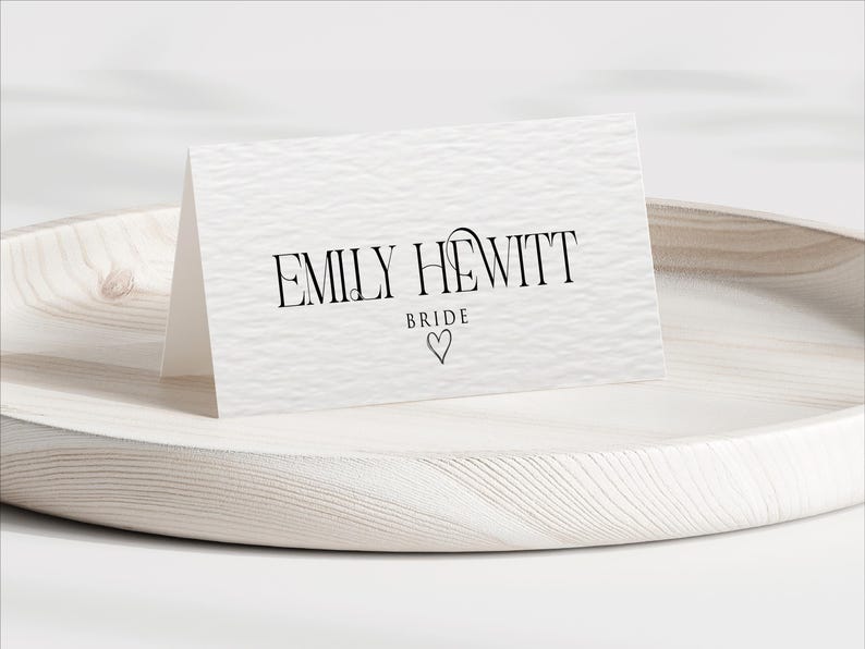 Pu&ograve; includere: Un biglietto bianco strutturato con il nome "EMILY HEWITT" in corsivo nero, con "BRIDE" sotto e un simbolo a forma di cuore. Il biglietto &egrave; esposto su un vassoio di legno chiaro.