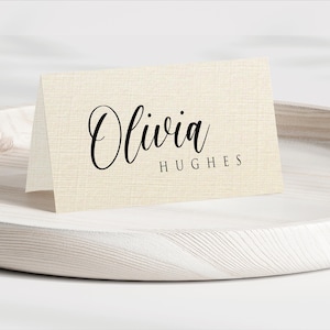 Puede incluir: Una tarjeta plegada con el nombre "Olivia" en elegante escritura negra y el apellido "Hughes" debajo. La tarjeta es de color beige claro y está sobre una bandeja de madera blanca.