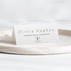Puede incluir: Una tarjeta de visita blanca con el nombre "Olivia Hughes" y el título "Senior Sales Executive" impresos en negro. La tarjeta tiene una superficie texturizada y el logotipo de Tidal Print en la parte inferior. La tarjeta se muestra en una bandeja de madera clara.