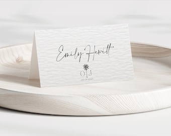 Puede incluir: Tarjeta blanca texturizada con el nombre "Emily Hewitt" en escritura cursiva, un icono de palmera, y las iniciales "O J" con la fecha "06.06.2023". La tarjeta se muestra en una bandeja de madera clara, para bodas o eventos especiales.