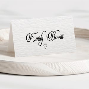Pu&ograve; includere: Un biglietto bianco piegato con il nome "Emily Hewitt" in elegante scrittura nera e un piccolo simbolo a forma di cuore. Il biglietto &egrave; appoggiato su un vassoio di legno chiaro con una superficie strutturata.