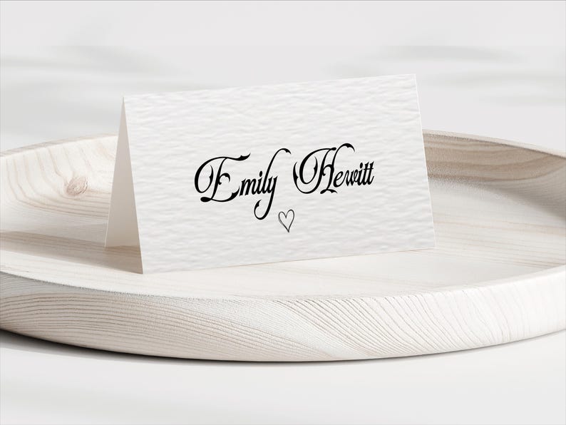 Pu&ograve; includere: Un biglietto bianco piegato con il nome "Emily Hewitt" in elegante scrittura nera e un piccolo simbolo a forma di cuore. Il biglietto &egrave; appoggiato su un vassoio di legno chiaro con una superficie strutturata.