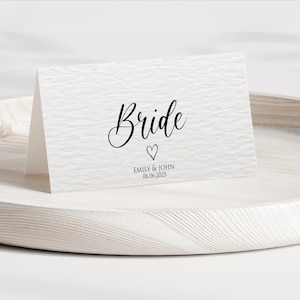 Pu&ograve; includere: Un biglietto bianco con la parola "Bride" in elegante scrittura nera, un cuore e i nomi "Emily & John" con la data "06.06.2025". Il biglietto &egrave; su un vassoio di legno chiaro.