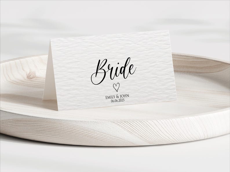 Pu&ograve; includere: Un biglietto bianco con la parola "Bride" in elegante scrittura nera, un cuore e i nomi "Emily & John" con la data "06.06.2025". Il biglietto &egrave; su un vassoio di legno chiaro.