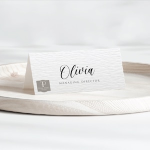 Puede incluir: Una tarjeta de lugar blanca texturizada con el nombre "Olivia" en escritura elegante y el título "Managing Director" en una fuente sencilla. El logotipo de Tidal Print está en la esquina inferior izquierda. La tarjeta descansa sobre una bandeja de madera clara.