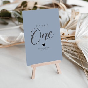Può includere: Un segnaposto blu chiaro con la parola "One" in elegante corsivo, un piccolo cuore e nomi con una data. Il segnaposto è esposto su un piccolo cavalletto di legno, ideale per un matrimonio o un evento.