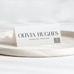Puede incluir: Una tarjeta de visita blanca y texturizada con el nombre "OLIVIA HUGHES" en negro, junto con "MANAGING DIRECTOR" debajo. Un logotipo gris está en la esquina inferior derecha. La tarjeta está sobre una bandeja de madera clara.