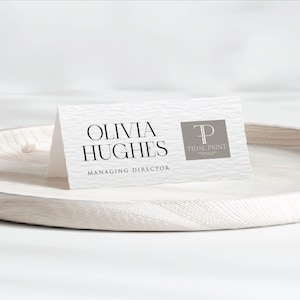 Puede incluir: Una tarjeta de visita blanca con el nombre "Olivia Hughes" y el título "Managing Director" impresos en negro. La tarjeta también presenta un cuadrado gris con el logotipo "Tidal Print". La tarjeta se muestra en una bandeja de madera blanca texturizada.