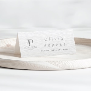 Puede incluir: Una tarjeta de visita blanca plegada con el nombre "Olivia Hughes" y el título "Senior Sales Executive" impresos en negro. La tarjeta también presenta el logotipo "Tidal Print". La tarjeta está sobre una bandeja de madera blanca.