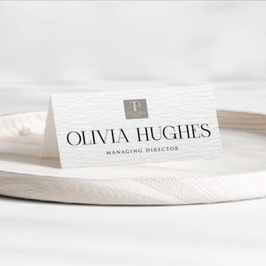 May include: A white business card with the name "OLIVIA HUGHES" in a serif font, and the title "MANAGING DIRECTOR" below. The card has a textured surface and a logo in the upper left corner. The card is on a white wooden tray.