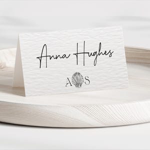 May include: White place card with black text that reads "Anna Hughes" and a seashell graphic with the letters "A" and "S" below it.