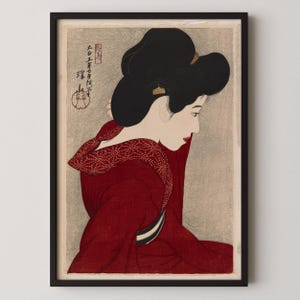 以下が含まれることがあります： 額装された日本の木版画で、横顔の女性が描かれています。彼女は模様の襟が付いた赤い着物を着ており、髪は伝統的なアップスタイルにまとめられています。プリントは、灰色の背景を持つ落ち着いた色合いです。