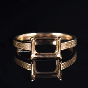 Könnte beinhalten: Ein goldfarbener Ring mit quadratischer Fassung, zur Aufnahme eines Edelsteins. Der Ring hat ein strukturiertes Band und eine polierte Oberfläche, die das Licht reflektiert. Der Ring wird auf einer reflektierenden Oberfläche präsentiert.