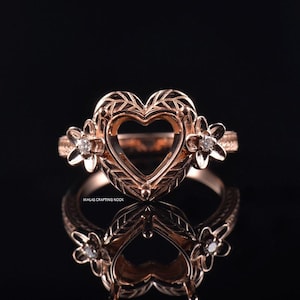 May include: Rose gold ring with a heart-shaped centre and floral accents. The ring features a heart-shaped setting with a decorative leaf pattern and small flower details on each side, each with a small diamond. The ring is reflected on a black surface.