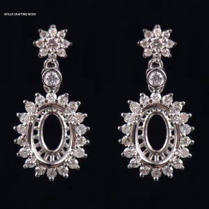 May include: A pair of silver dangle earrings with a round, oval-shaped setting for a gemstone. Each earring features a cluster of small, clear stones at the top and a larger cluster of stones surrounding the oval setting.