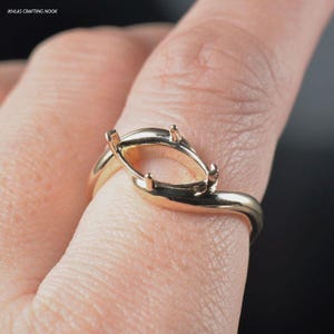 Puede incluir: Un anillo de oro con una banda retorcida y un engaste ovalado para una piedra preciosa. El anillo se muestra en un dedo.