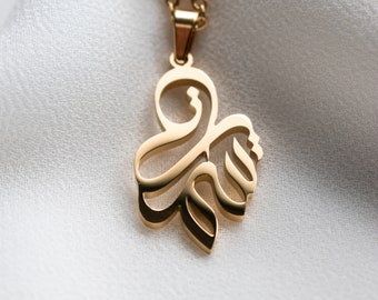 May include: A gold-toned necklace with a pendant shaped like a stylized Arabic letter. The pendant is intricate and detailed, with a delicate design.