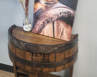 May include: A half-barrel table with a dark wood finish and metal bands. A canvas print of a whiskey glass on a barrel sits on top. A small glass bottle with dried flowers is also on the table.