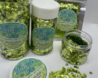 May include: Several containers of holographic leaf glitter in various sizes. The glitter is lime green and shaped like cannabis leaves. The containers include jars, a bag, and a small tub. The labels read "Loud OG" and "Holographic Leaf Glitter."