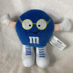 May include: Blue plush M&M character wearing glasses and white shoes with blue stripes. The character has a smiling face and the text "Brilliant Blue" on the tag.