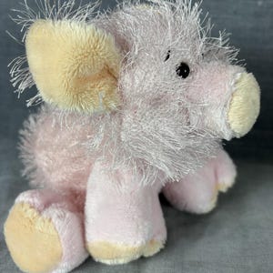 Peut inclure: Une peluche en forme de cochon avec une fourrure rose clair et de longues fibres blanches ébouriffées sur la tête et les oreilles. Le museau, l'intérieur des oreilles et les pieds sont d'un jaune pâle contrastant. Deux petits yeux noirs.
