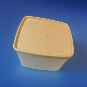 Peut inclure: Un récipient Tupperware carré, blanc cassé, avec un couvercle assorti. Le couvercle porte le logo Tupperware et une empreinte circulaire au centre. Le récipient est posé sur un fond bleu.