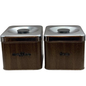 May include: Two brown woodgrain metal canisters with silver lids. The lids have a small black knob on top. The canister on the left has the word "Coffee" printed on the front. The canister on the right has the word "Tea" printed on the front.