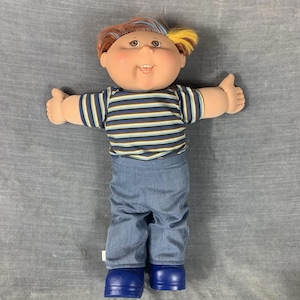 Peut inclure: Poupée Cabbage Patch Kid vintage avec un visage rond, des yeux bruns et une expression souriante. La poupée a des cheveux bruns et jaunes, une chemise rayée, un jean bleu et des chaussures bleues. La poupée est debout, les bras tendus.